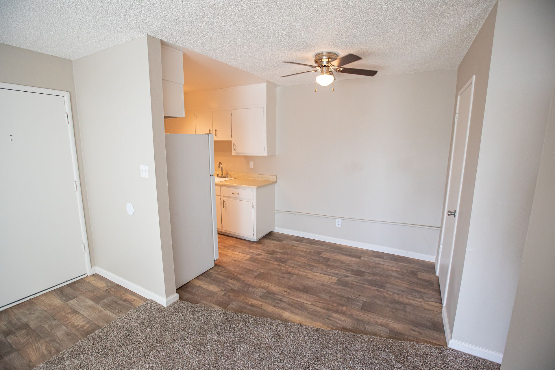 Empty apartment living area with carpet, wood-look flooring, kitchen nook, and ceiling fan