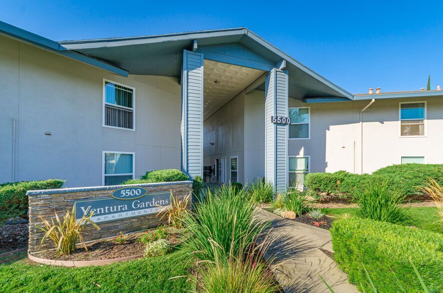 Modern apartment entrance with sign, walkway, and landscaped greenery under a blue sky