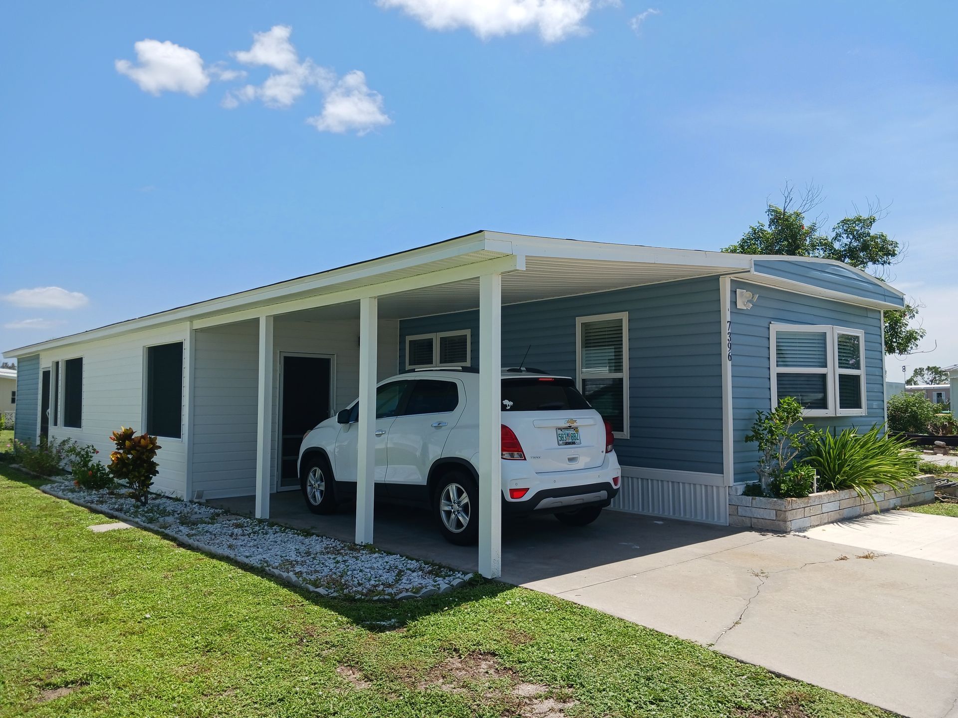 Blue and white mobile home with attached carport, white SUV parked underneath, sunny day. Blue and white mobile home with attached carport, white SUV parked underneath, sunny day.