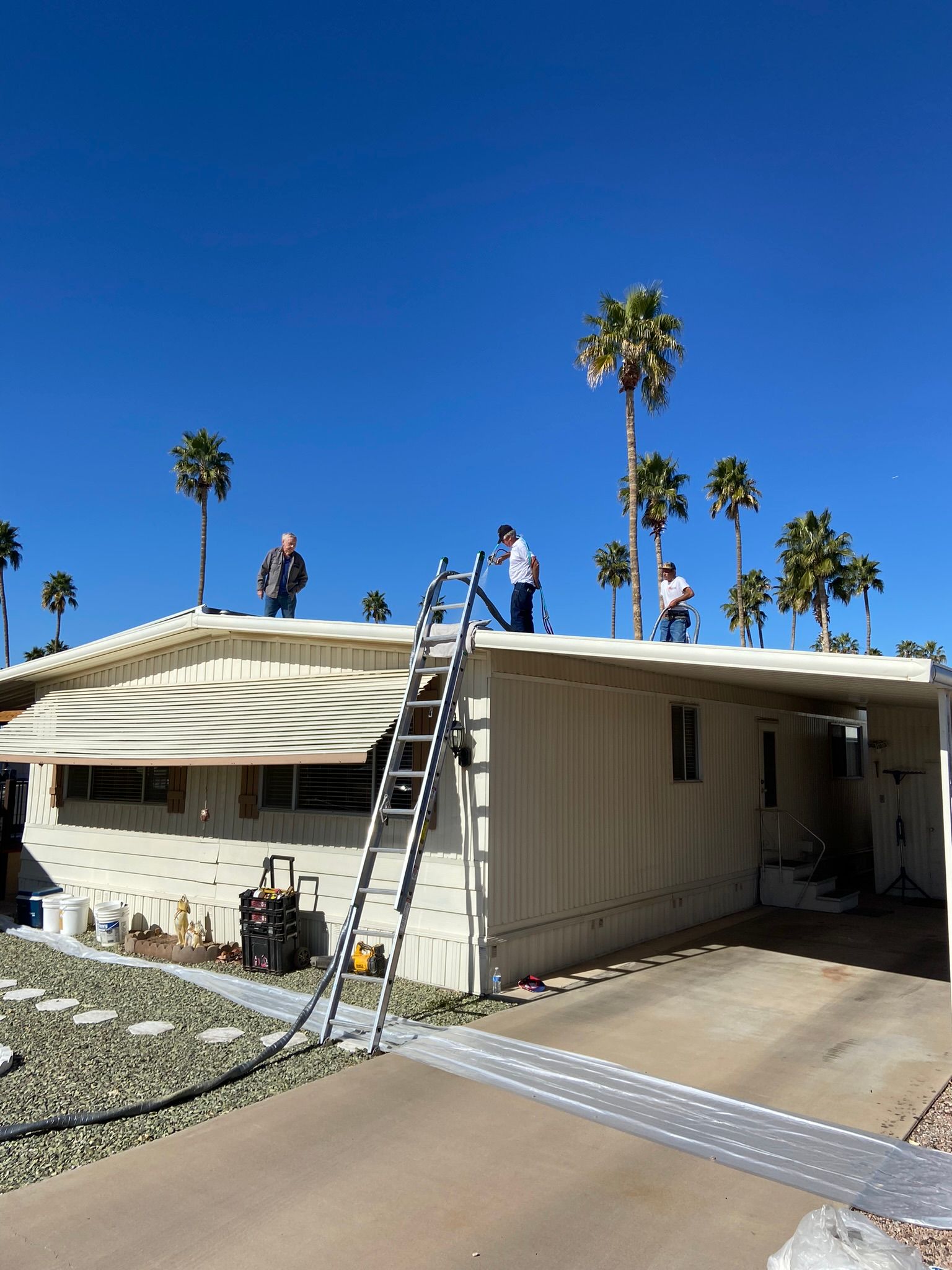 Workers repair a roof with ladders on a single-story house under a clear blue sky.