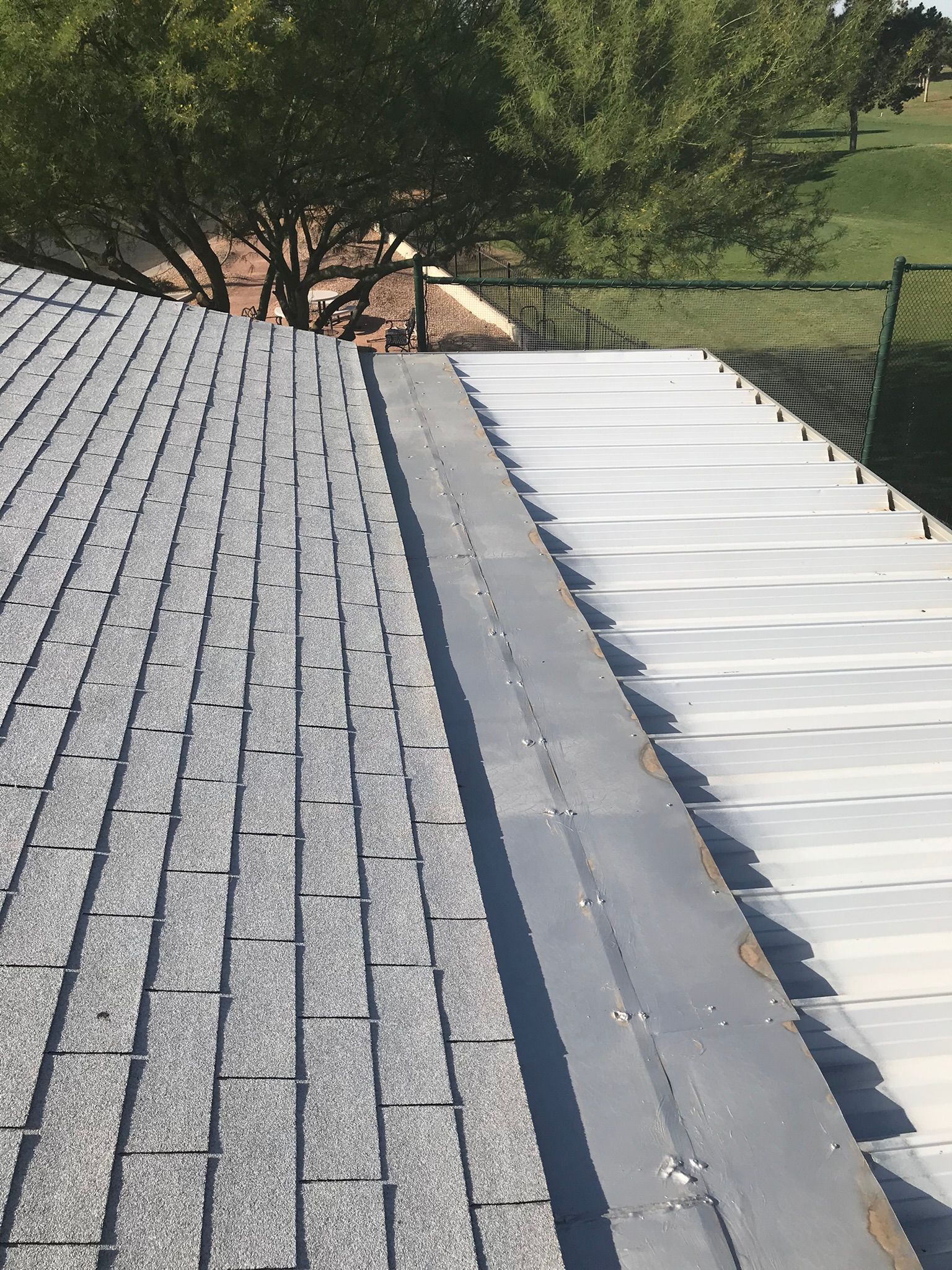 Rooftop with gray shingles beside a white corrugated roof edge and trees in the background