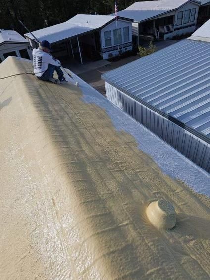 Worker on a sloped roof beside a metal-roofed building, applying yellow roof coating.