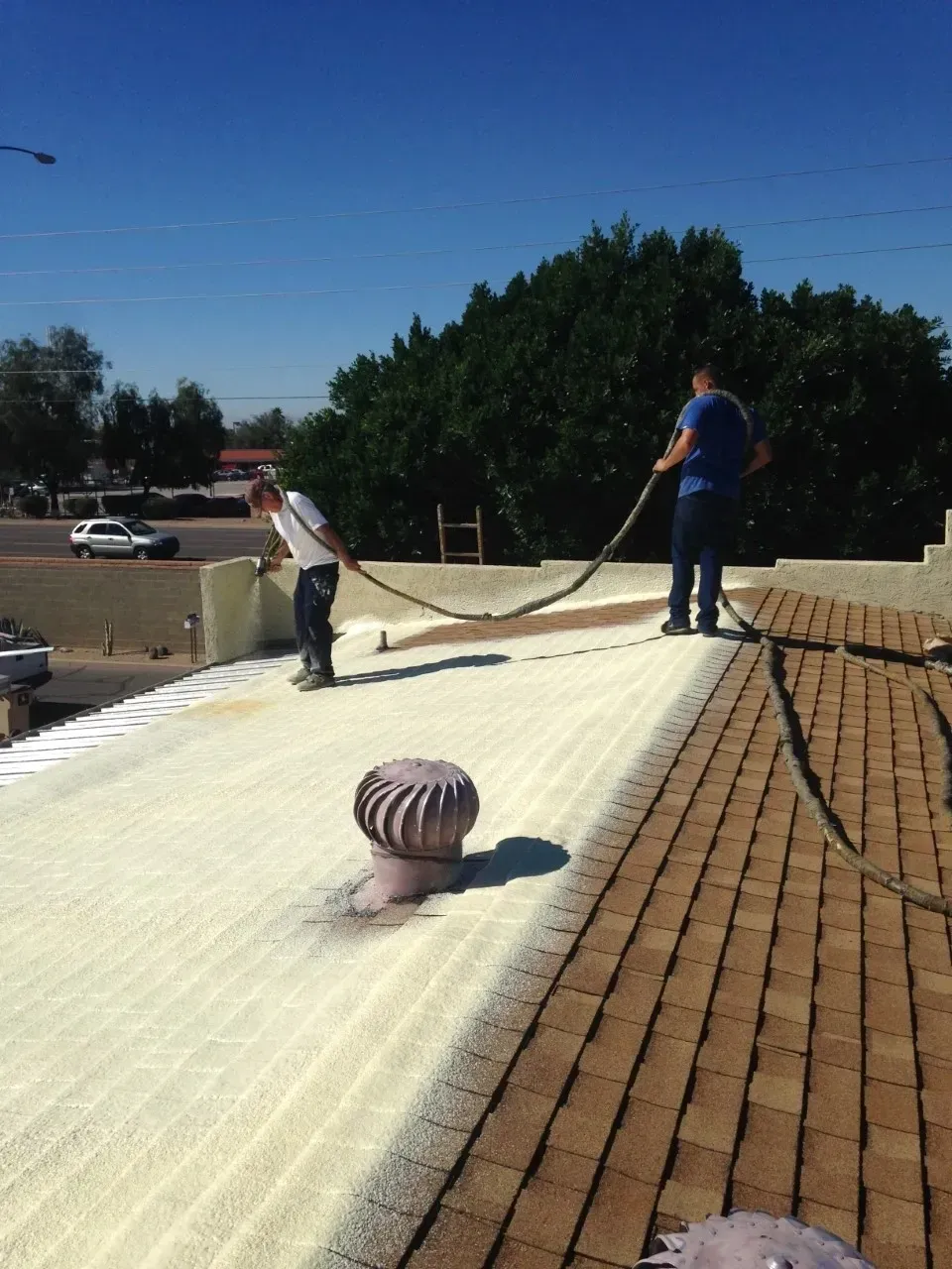 Two workers spread roofing material on a flat roof under a clear blue sky.