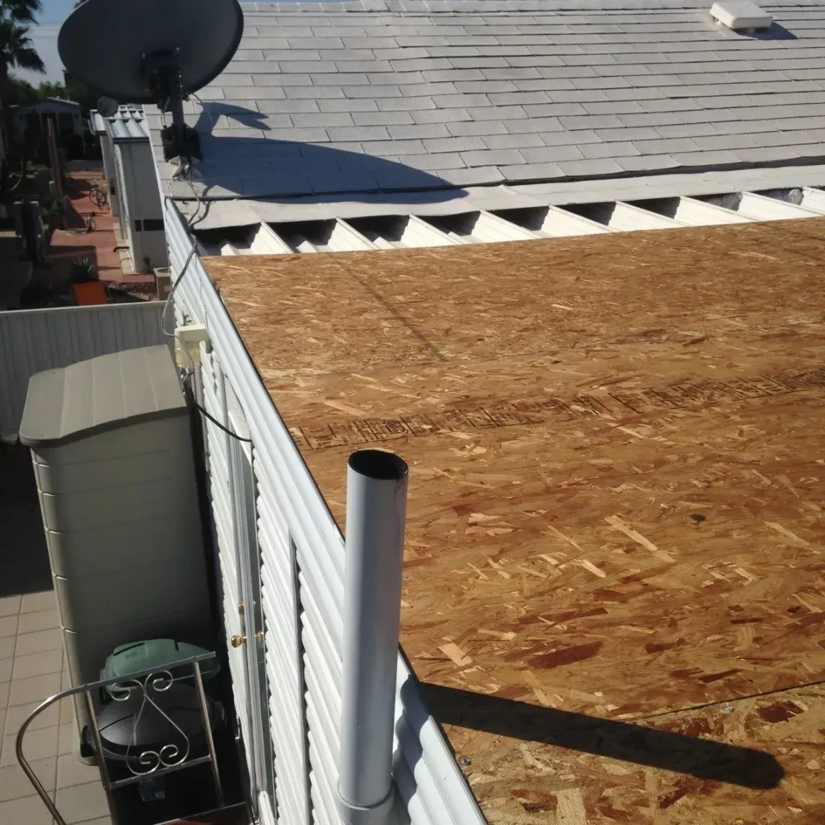 Rooftop with plywood sheathing, white trim, satellite dish, and a vent pipe beside a neighboring building.