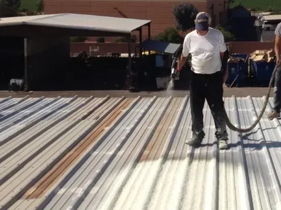 Worker standing on a corrugated metal roof, holding a hose in an outdoor construction area.