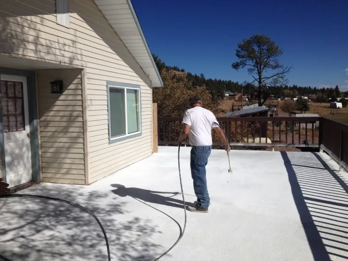 Person walking on a sunlit concrete patio beside a beige house, holding a hose with mountains in the background