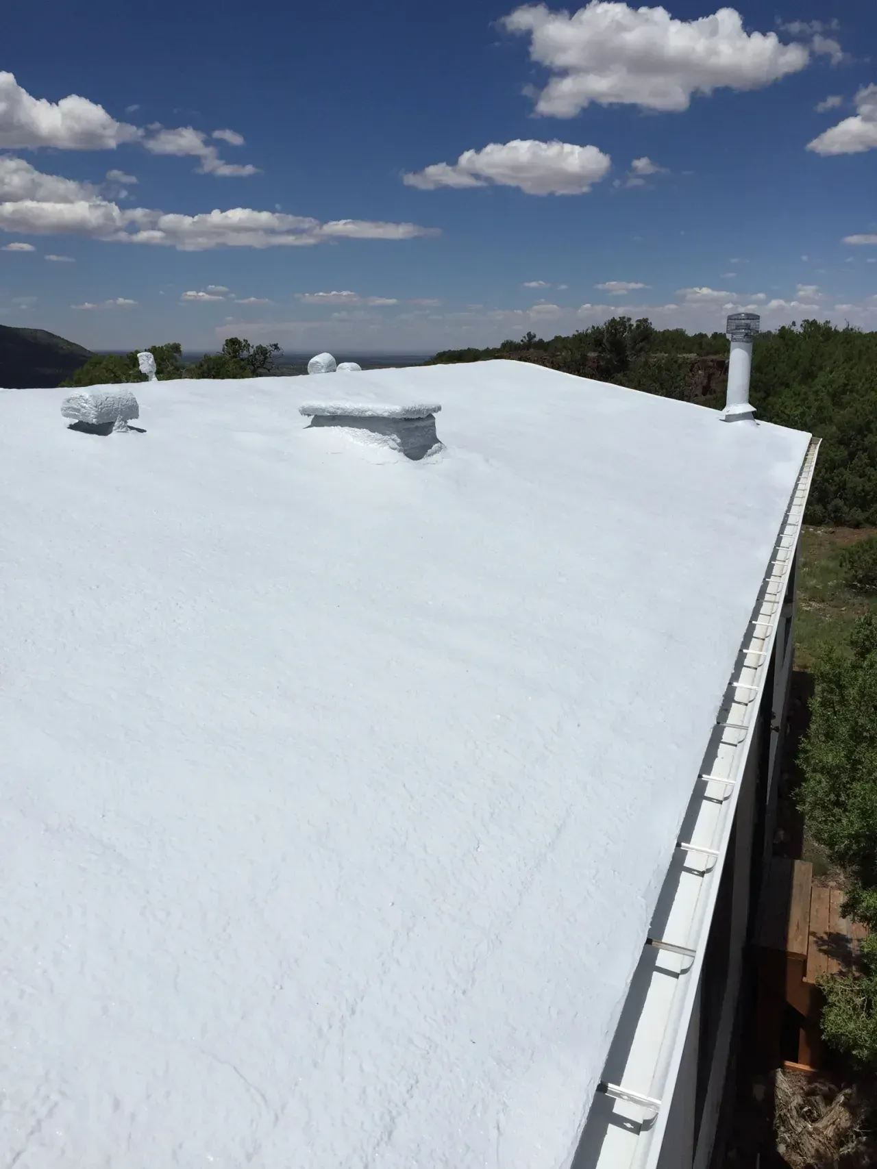 Snow-covered white roof under a blue sky with clouds, with a vent pipe and distant trees.