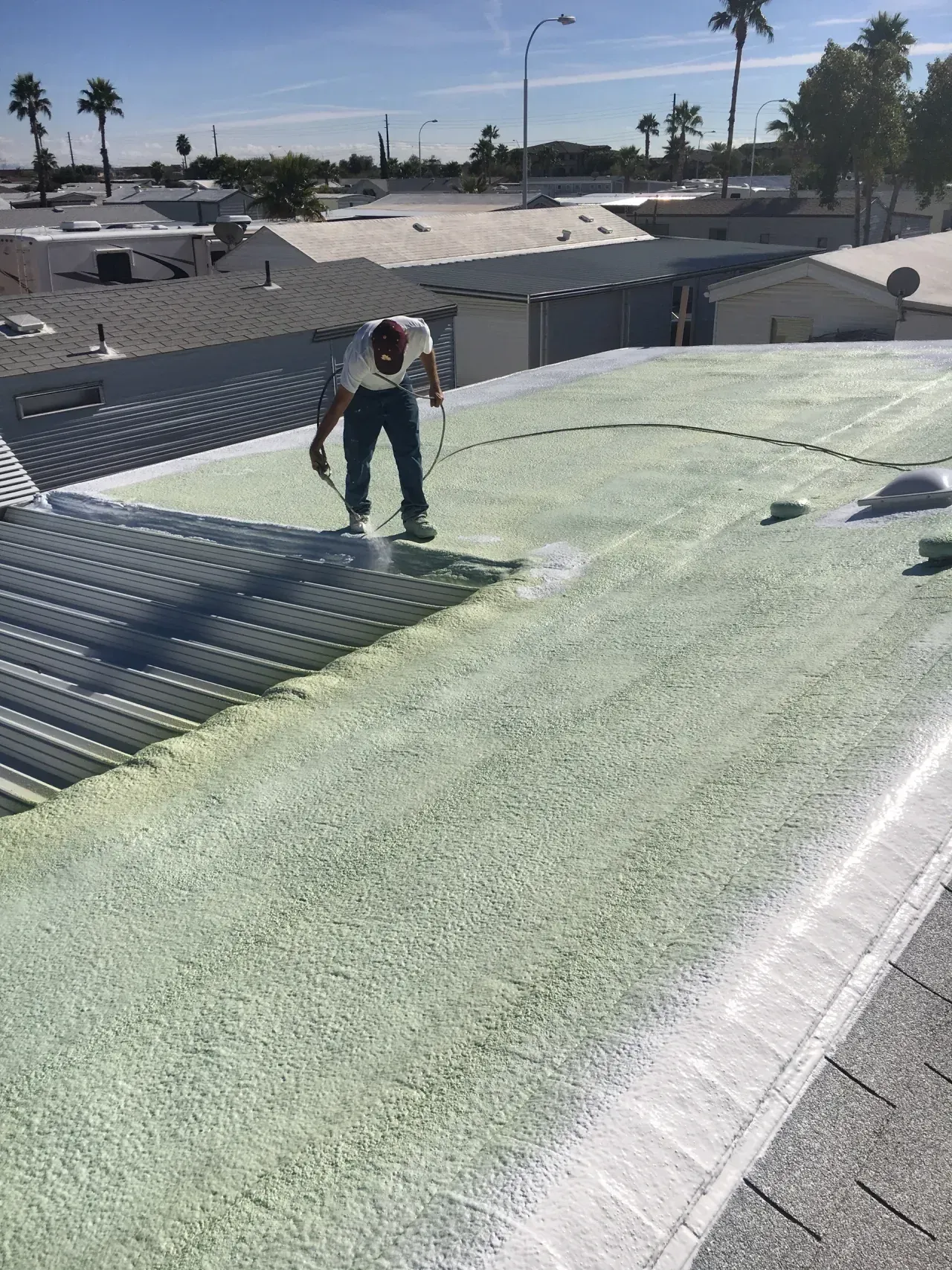 Worker applying green roof coating on a sloped rooftop near palm trees and houses