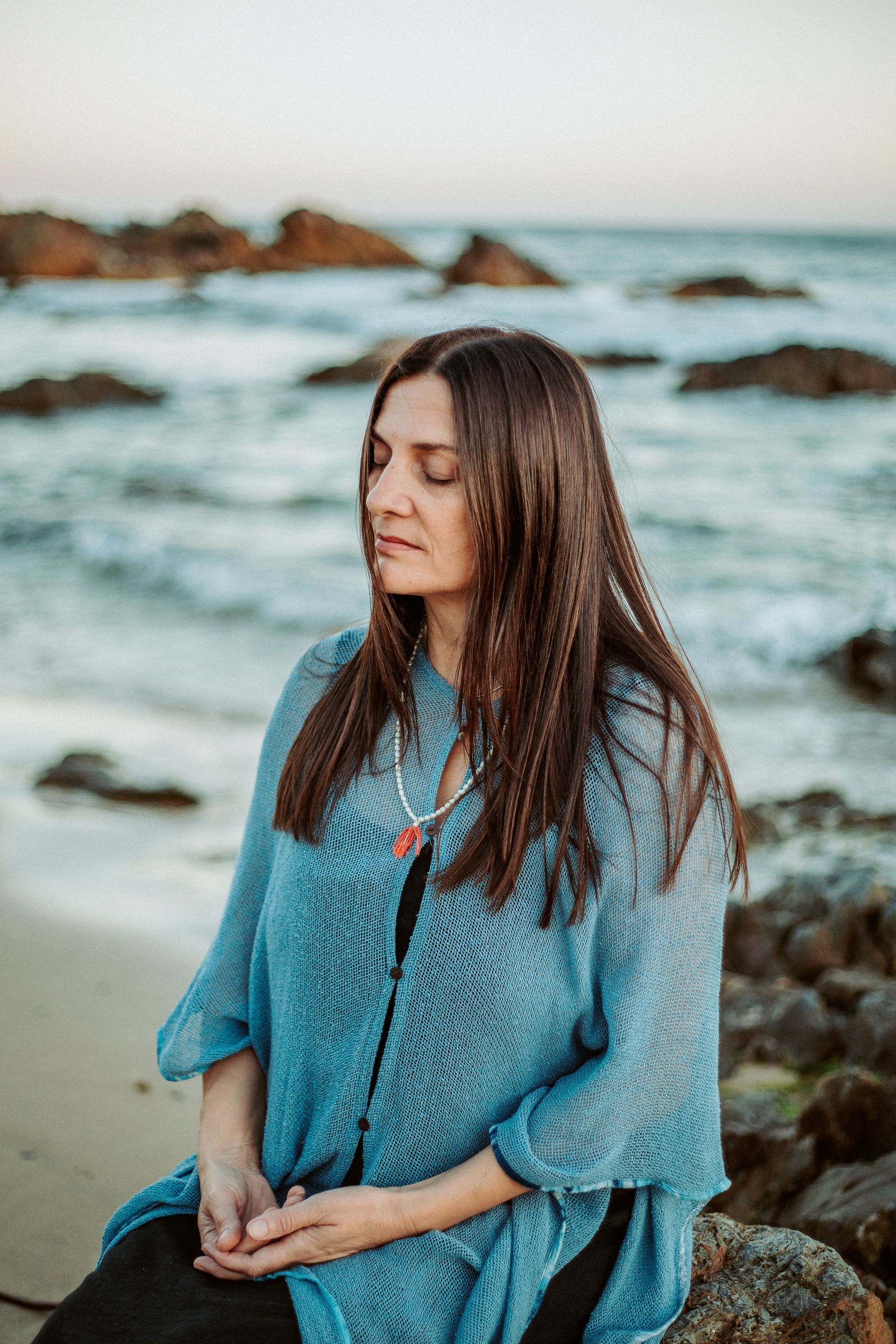 A person sits calmly with eyes closed on a rocky beach by the ocean — Vedic Meditation on the Coffs Coast, NSW