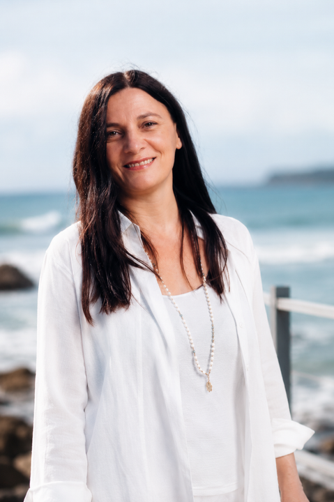 Woman with long dark hair smiles outside, wearing white shirt and necklace; ocean background.