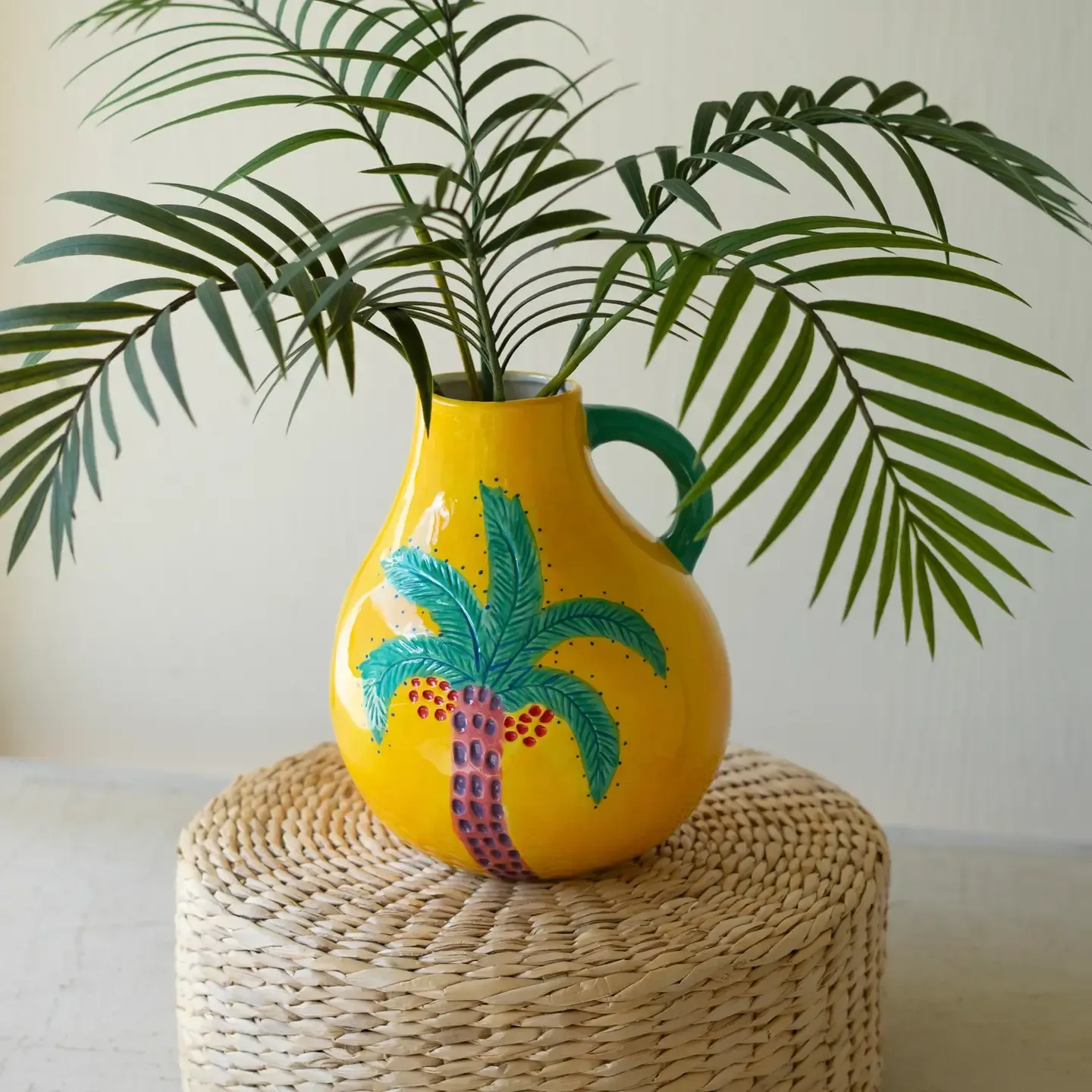 Yellow vase with palm design holding green fronds on a woven stool against a plain wall