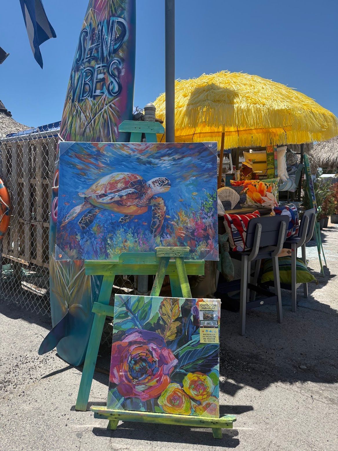 Outdoor art display with colorful seascape paintings and a bright yellow umbrella under a clear blue sky.