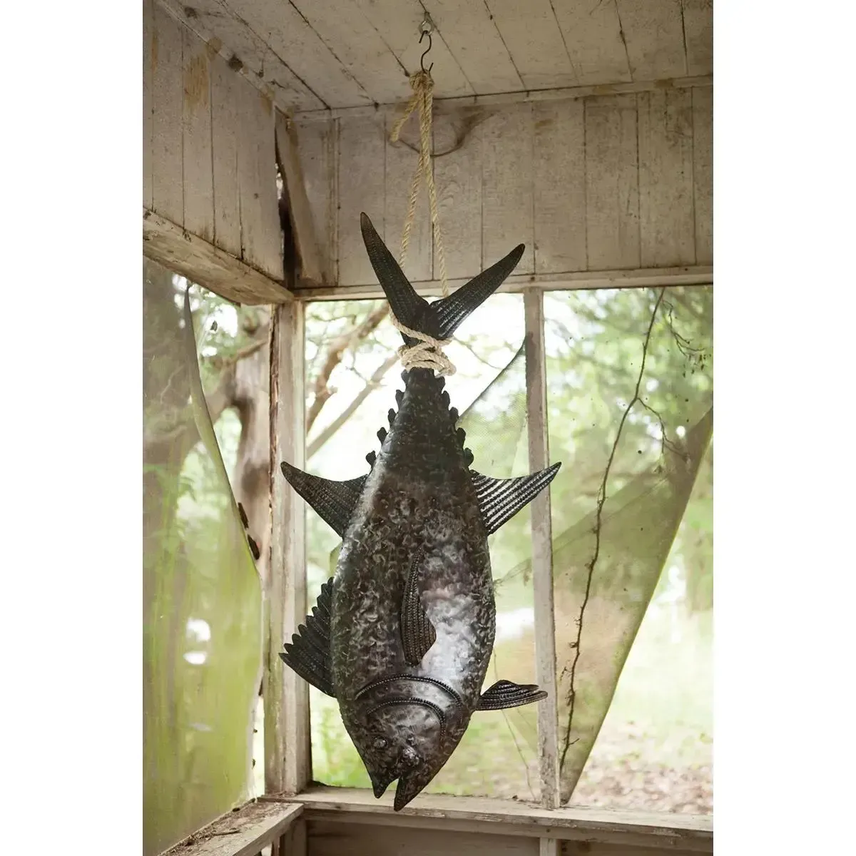 A large fish hangs upside down from a hook inside a rustic porch or shed.