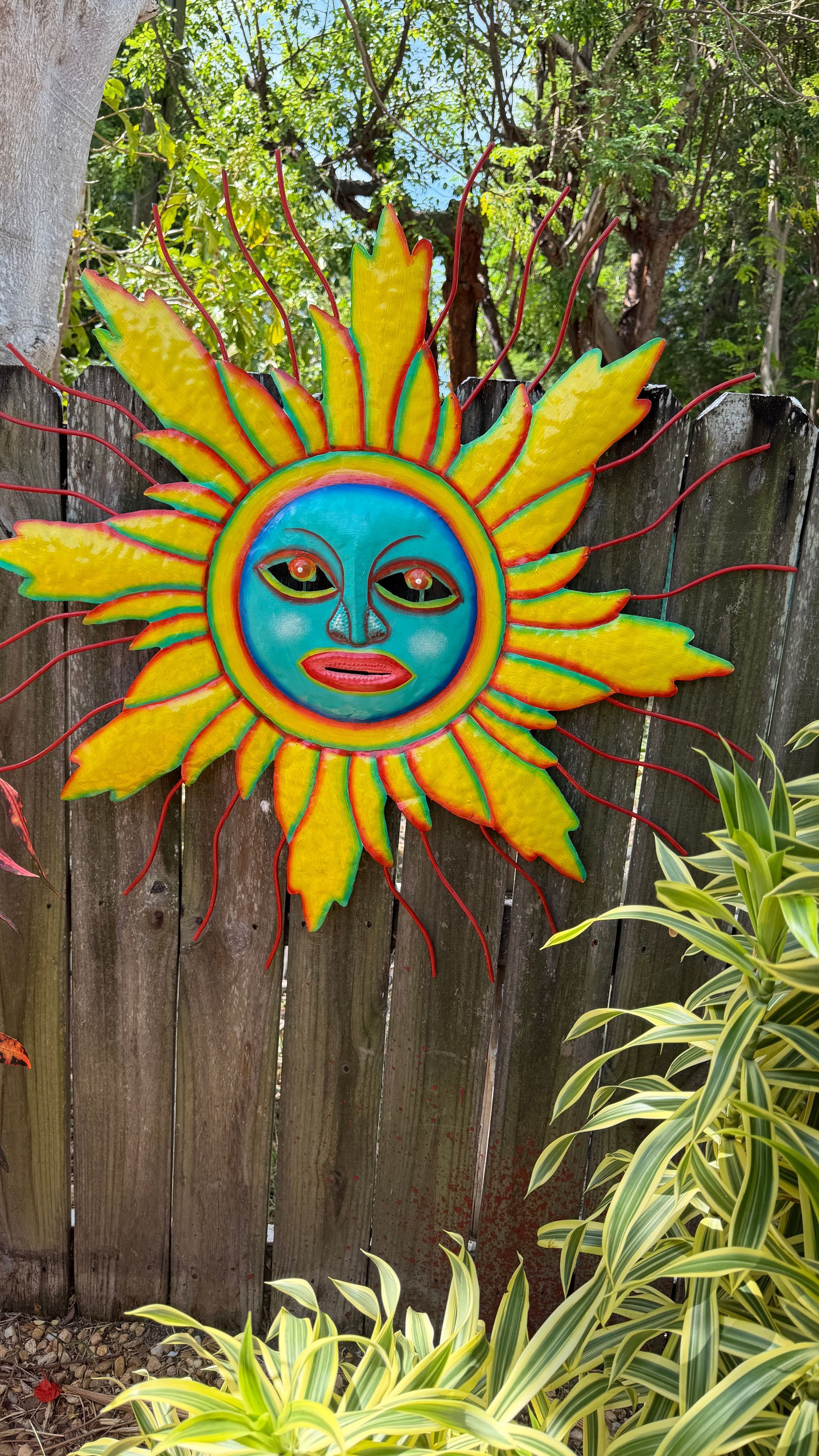 Colorful sun face wall art hanging on a wooden fence in a garden with green plants nearby