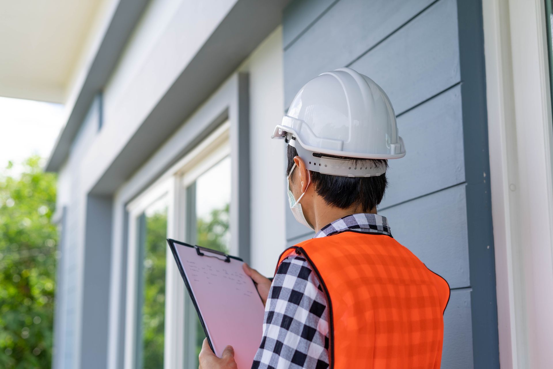 A man wearing a hard hat and an orange vest is holding a clipboard in front of a building.
