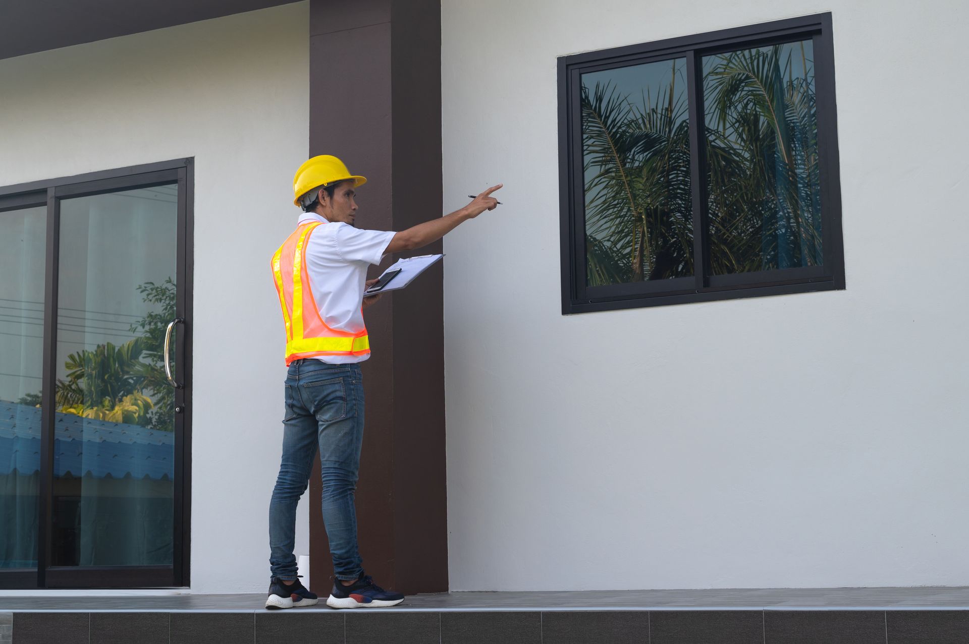A man wearing a hard hat and an orange vest is holding a clipboard in front of a building.