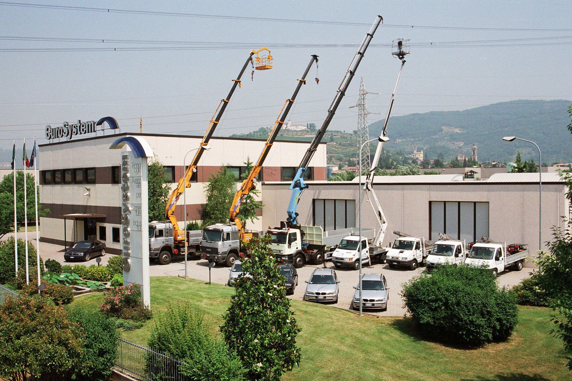 Gru da costruzione accanto a un edificio e auto parcheggiate sul ciglio erboso della strada, con colline sullo sfondo.