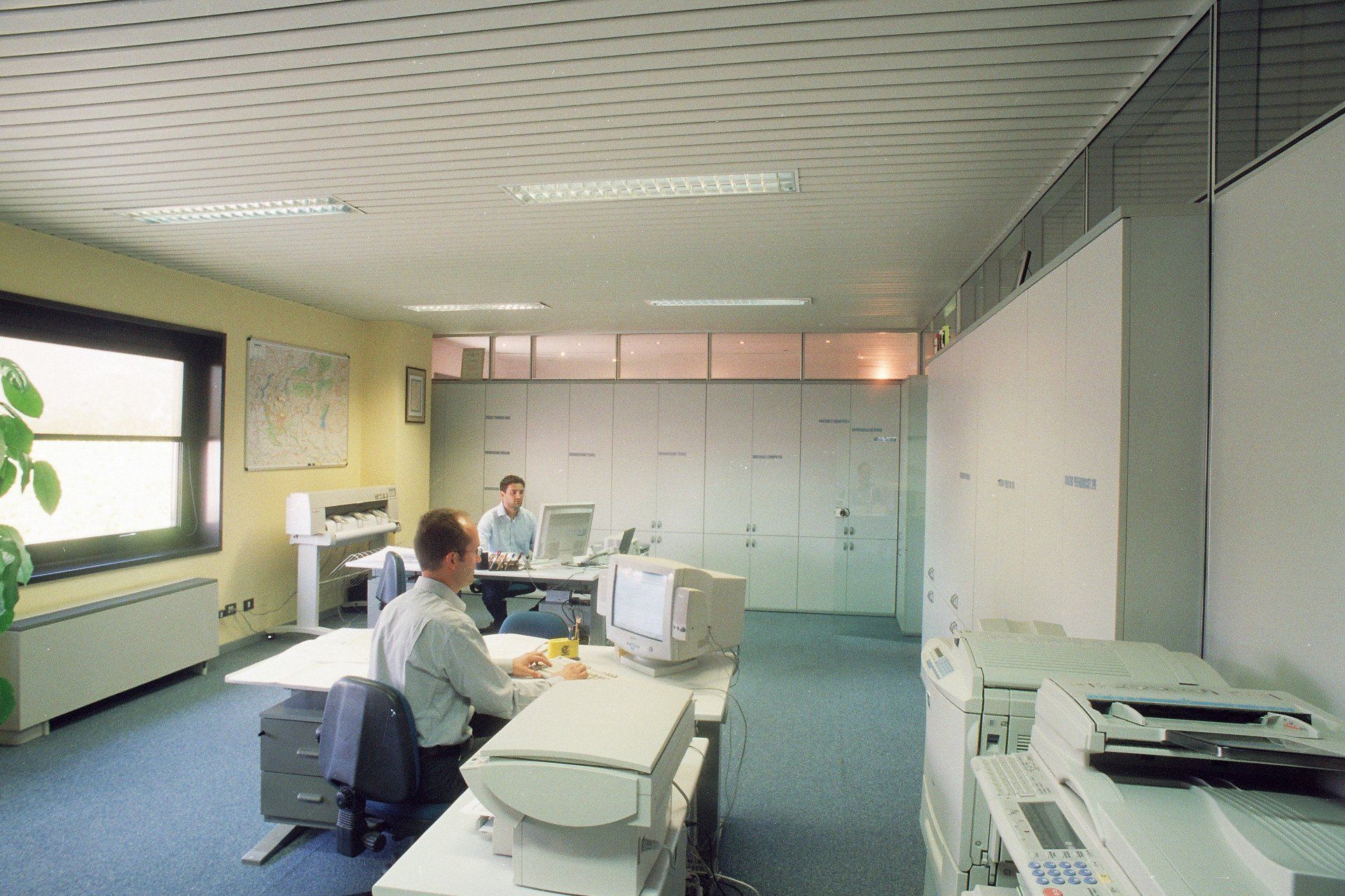 Postazione di lavoro in ufficio con due persone sedute a scrivanie, computer e schedari, illuminata da una luce fluorescente intensa.