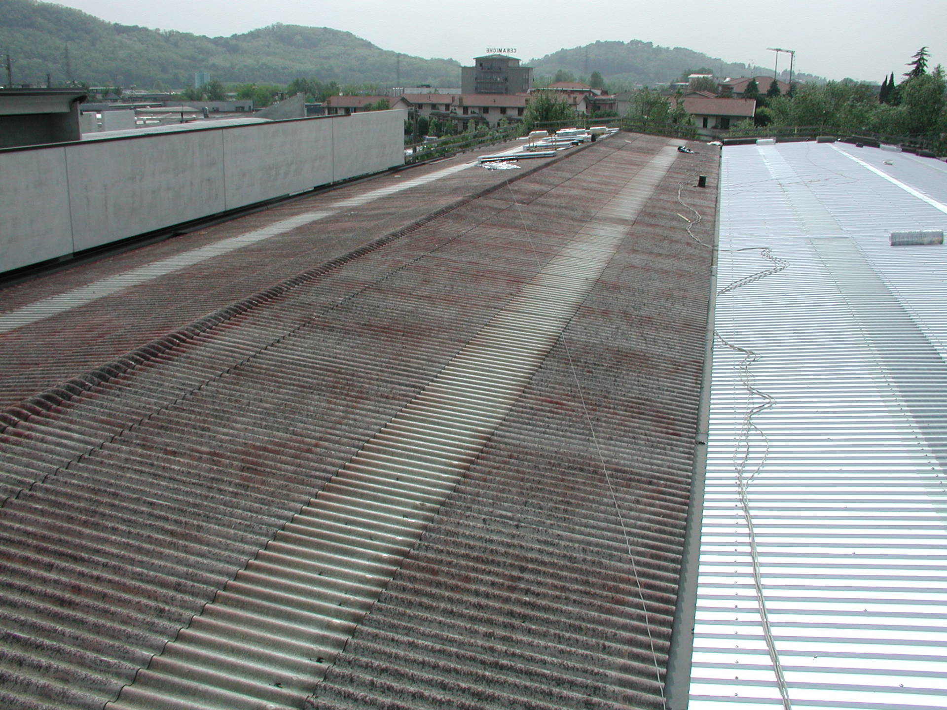 Tetto con file di pannelli metallici arrugginiti accanto a un tetto bianco, con colline ed edifici in lontananza.