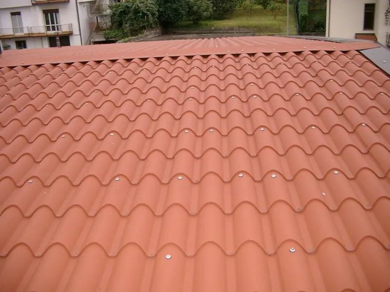 Tetto in tegole di terracotta con motivo ondulato, visto dall'alto, con edifici e alberi sullo sfondo.