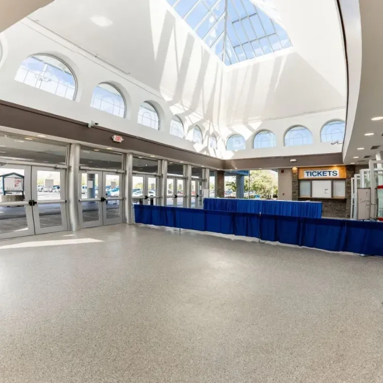 Spacious, brightly lit lobby with a high atrium ceiling, glass windows, tiled floor, and a blue-draped ticket counter.