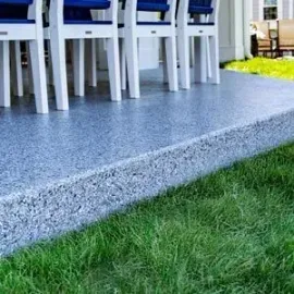 White chairs with blue cushions sit on a grey speckled patio overlooking a green lawn.
