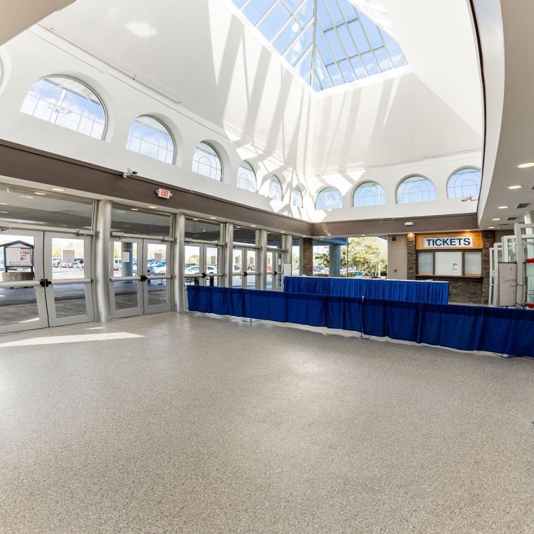 Spacious, brightly lit lobby with a high atrium ceiling, glass windows, tiled floor, and a blue-draped ticket counter.