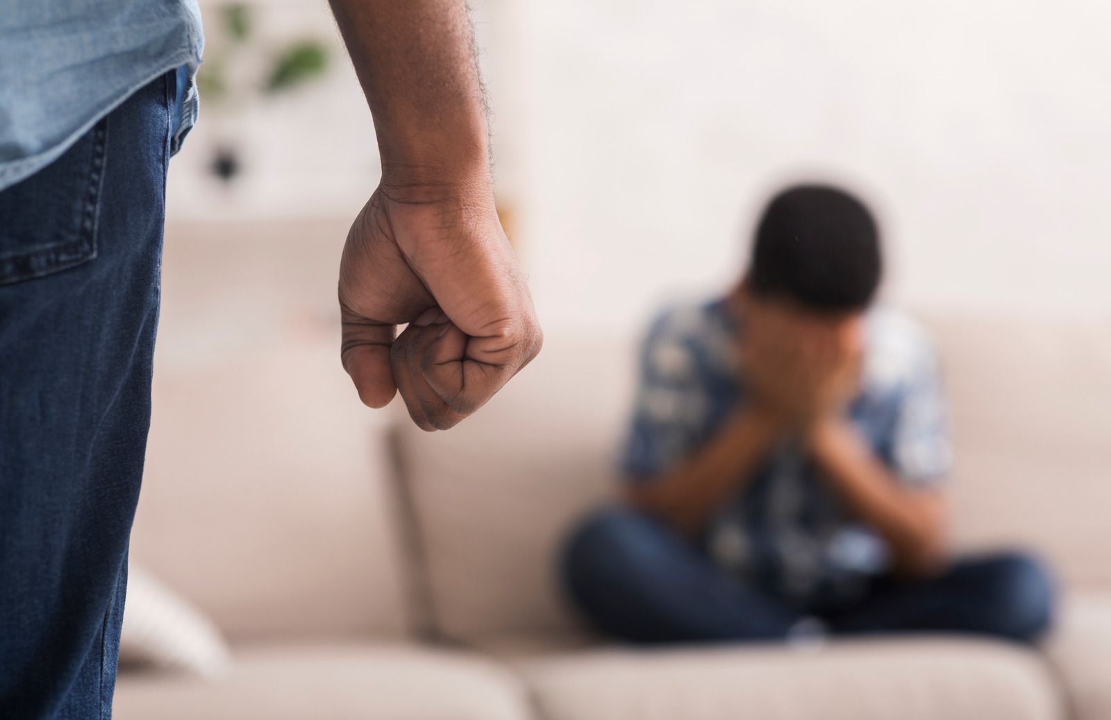 Clenched fist in foreground, with a blurred person sitting on a couch and covering their face in the background.