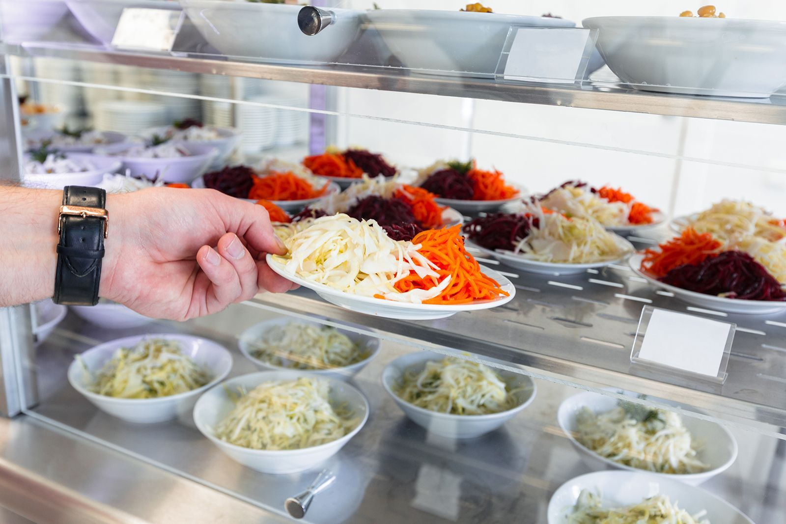 Selección manual de alimentos de una vitrina de cafetería. Se ven varias ensaladas en tazones y platos.