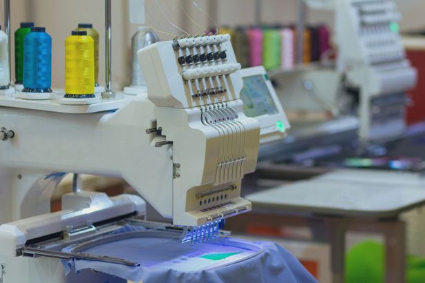 Una macchina da ricamo industriale cuce un disegno su un tessuto blu con rocchetti di filo colorati sullo sfondo.