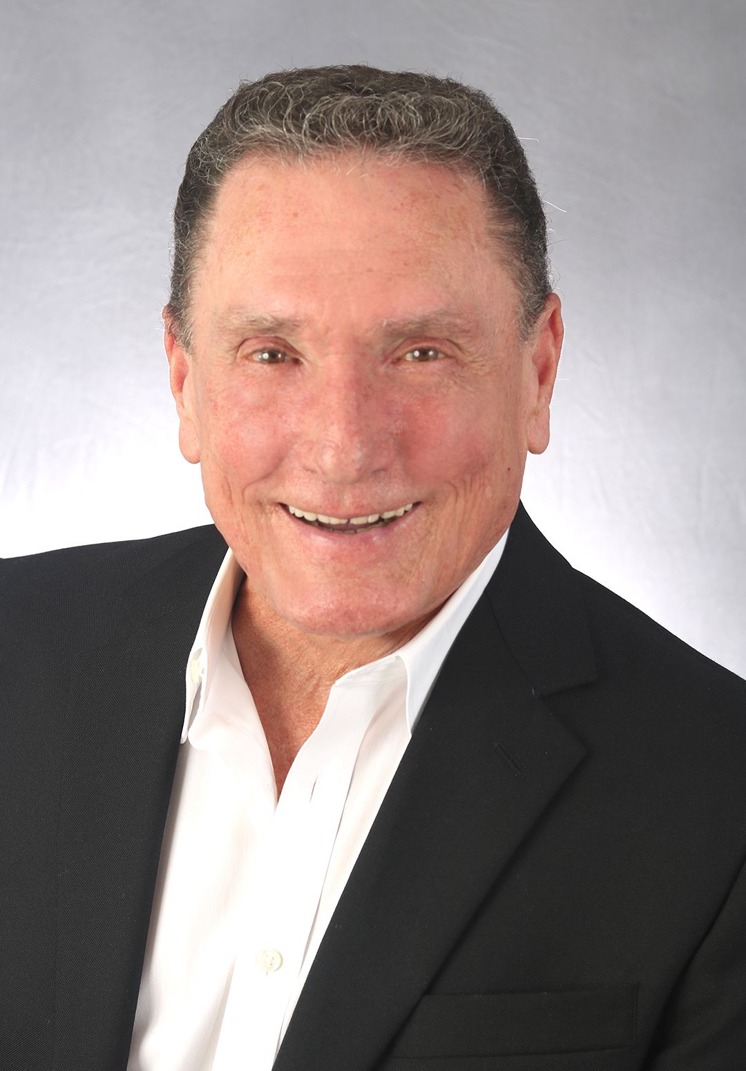 Man in a black suit and white shirt smiles at the camera against a gray background.