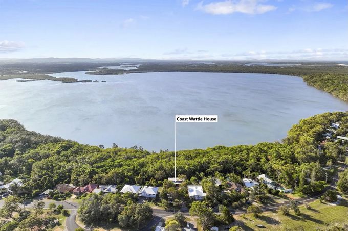 An aerial view of homes near a large lake, surrounded by trees under a bright blue sky. The text says "Coast Wattle House".