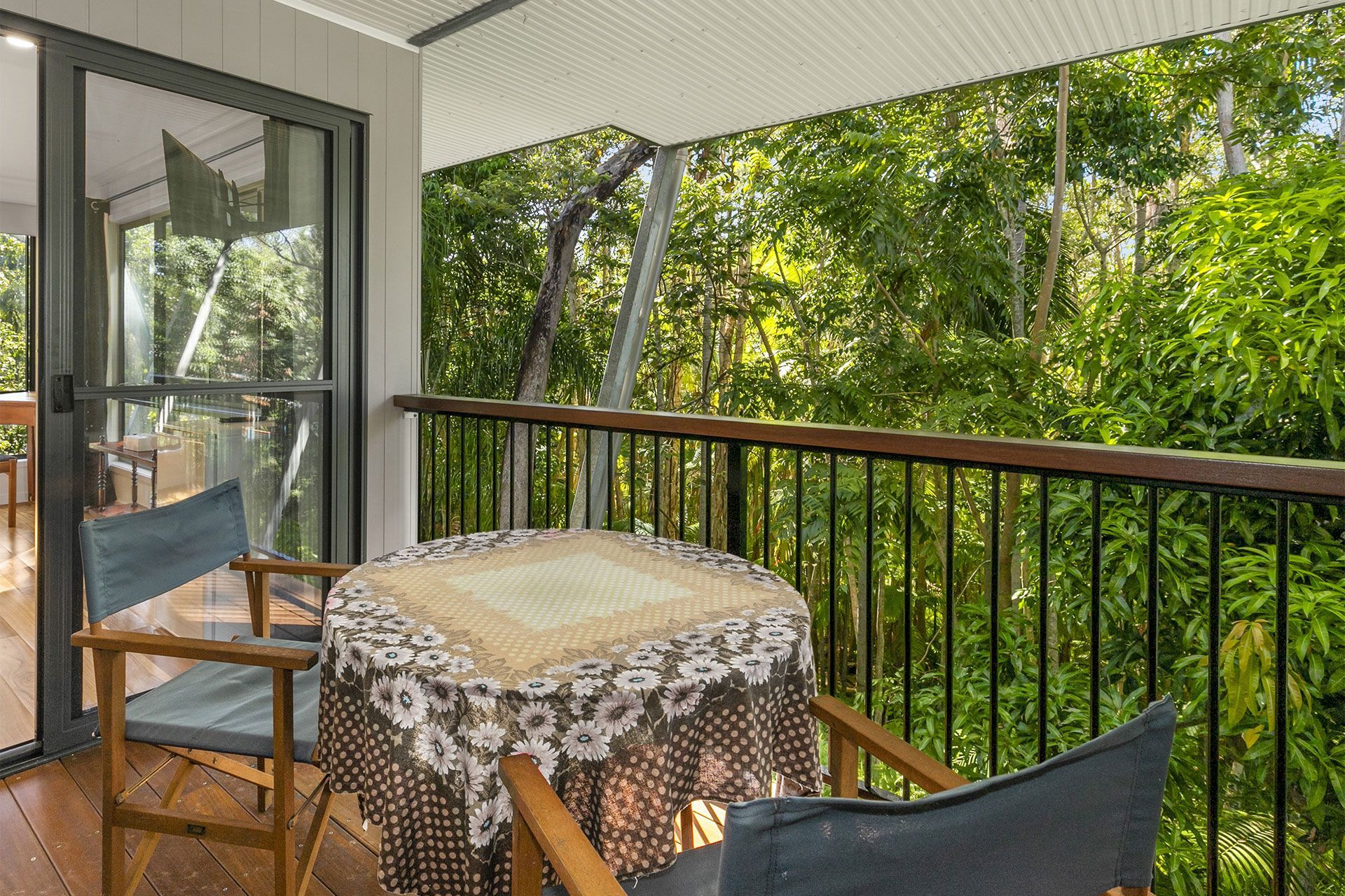 A balcony with a table and two chairs, overlooking lush green trees. The table is covered with a floral tablecloth.