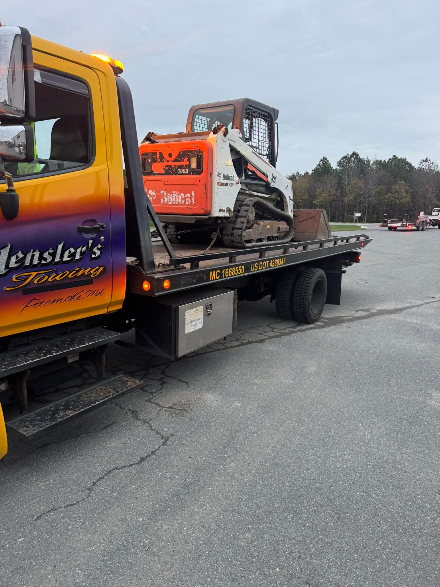 Yellow tow truck with a Bobcat skid steer on its flatbed. Outdoors, overcast.