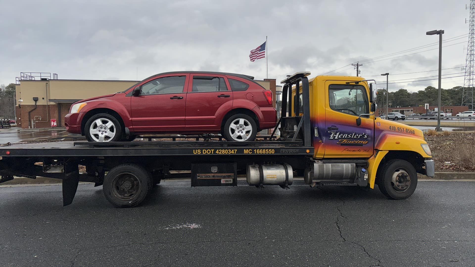 A red car being towed on a yellow and black tow truck. American flag in background.