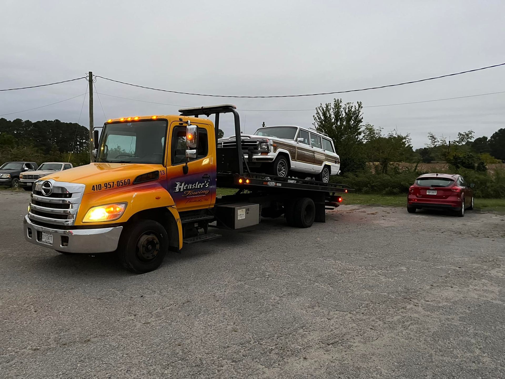 Yellow tow truck carrying a white vintage SUV on a gravel lot; red car nearby.