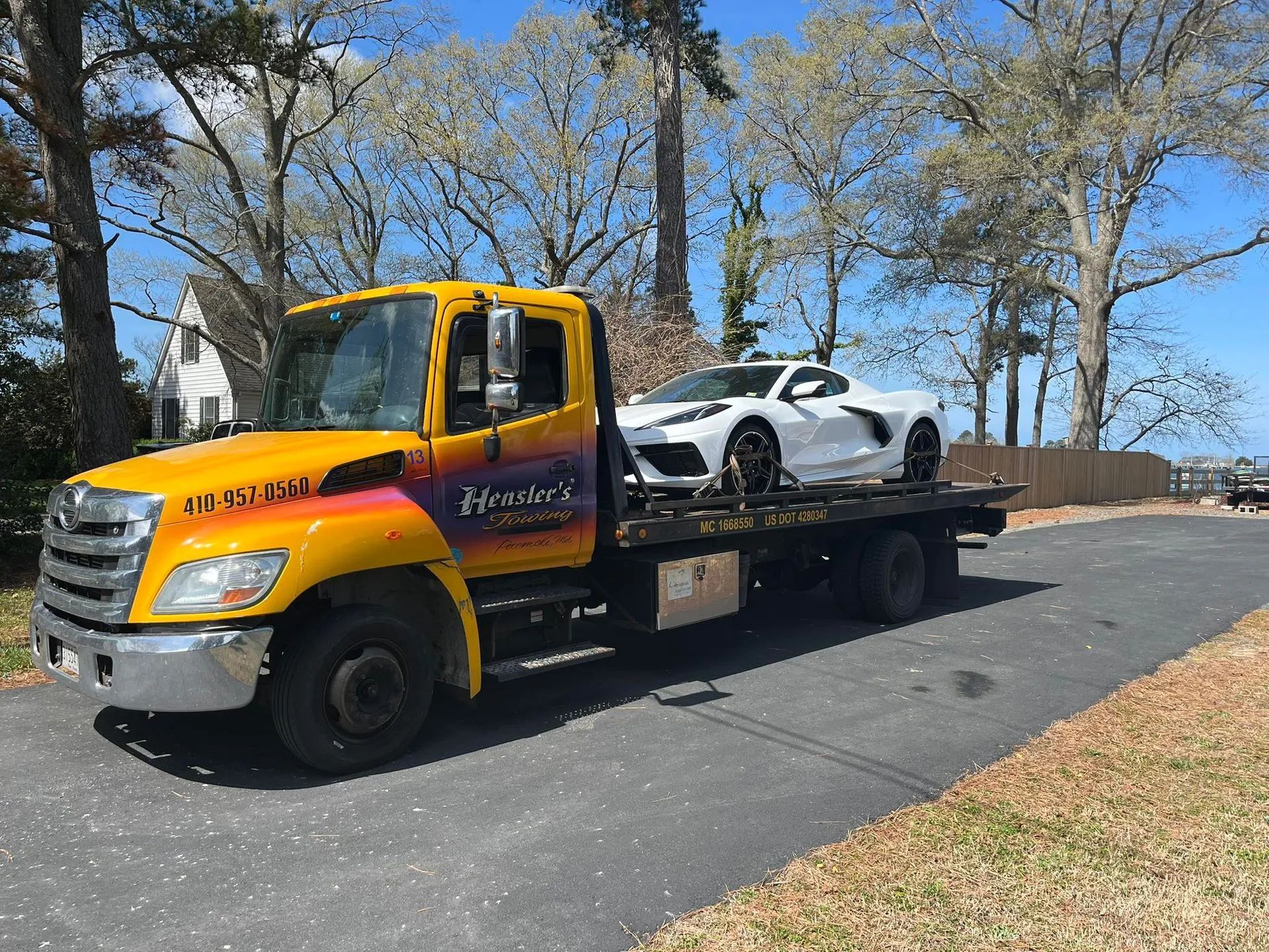 Yellow tow truck carrying a white sports car on a flatbed in a roadside lot under trees