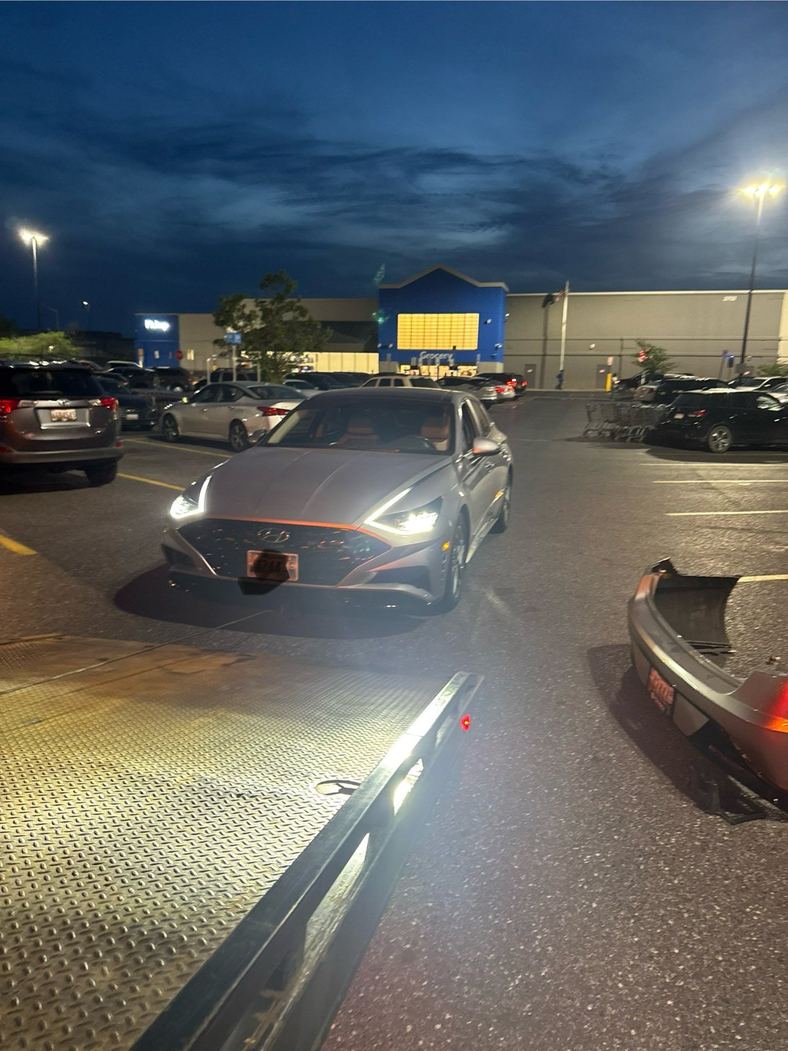 Silver car in a parking lot, headlights on, with a Walmart store in the background at dusk.