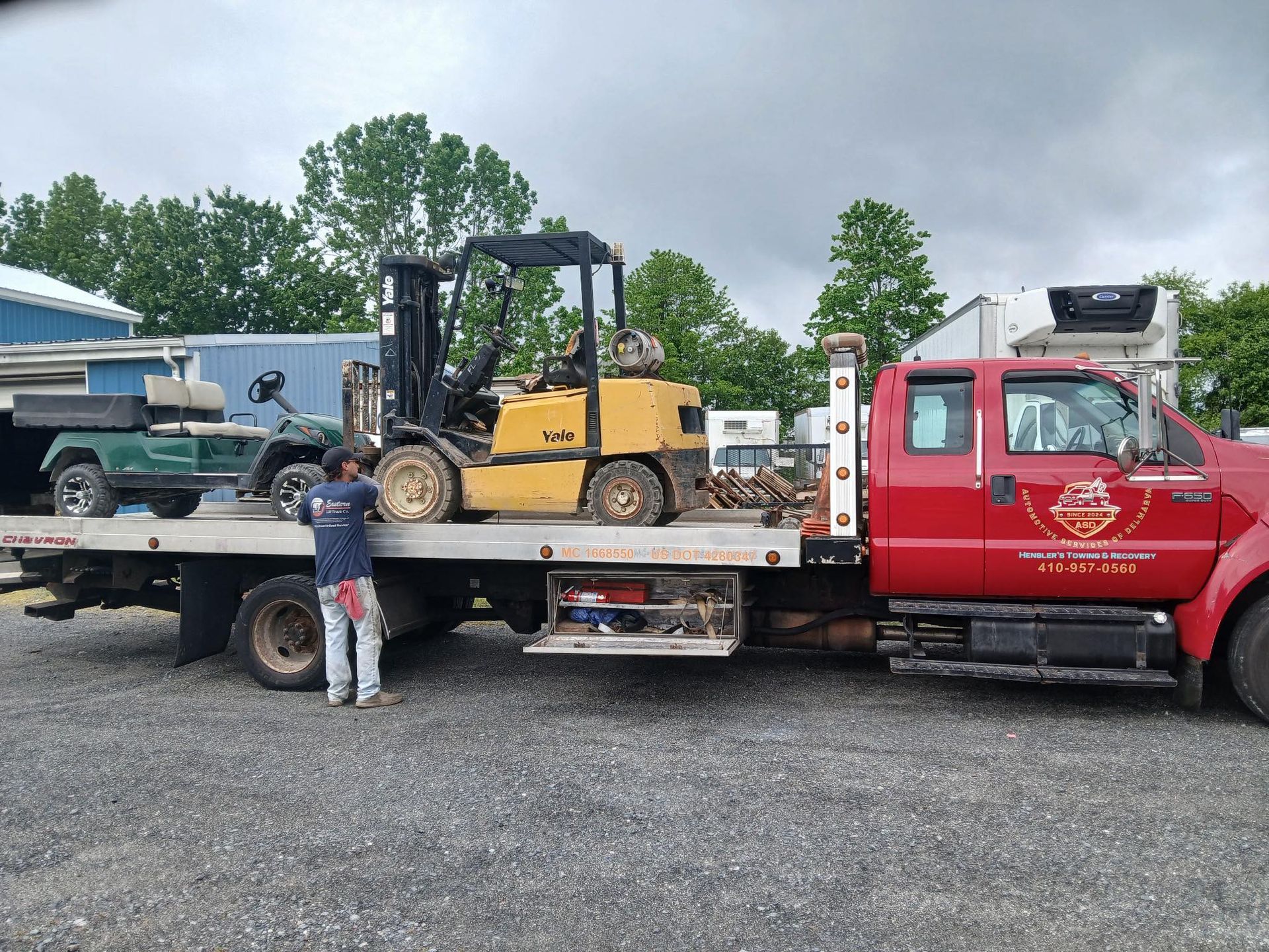 Forklift loaded onto a red flatbed tow truck by a man in a blue shirt.