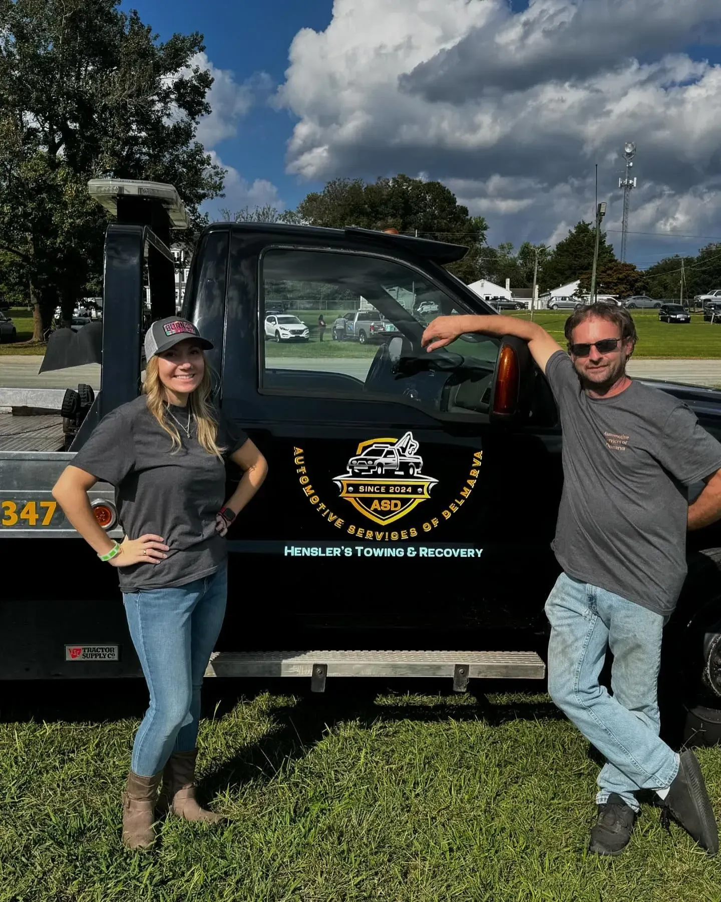 Two people by a black tow truck. One leans on the truck, the other has hands on hips. Logo on the side.