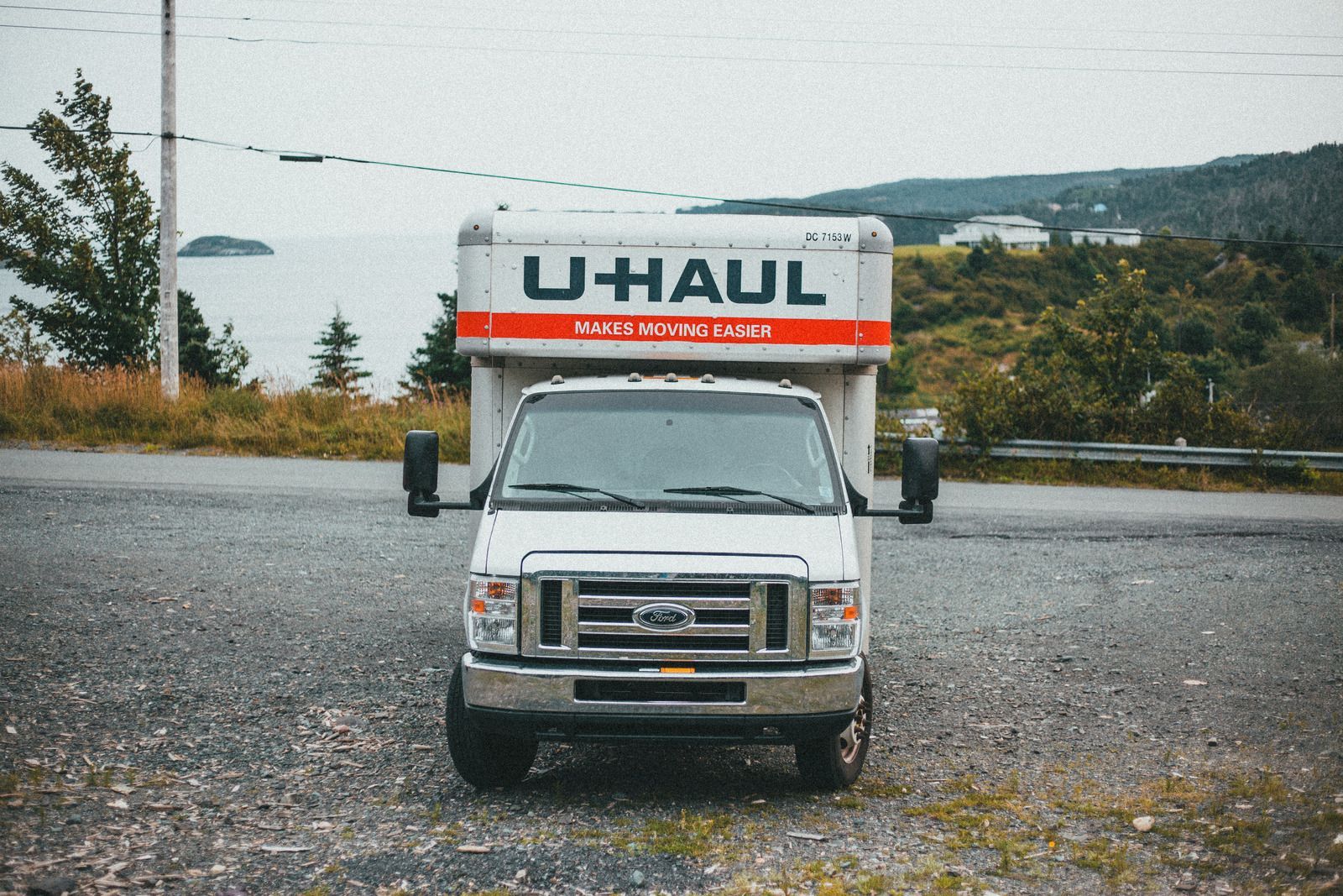 White U-Haul truck and trailer parked on a gravel road, overlooking a body of water and distant hills.