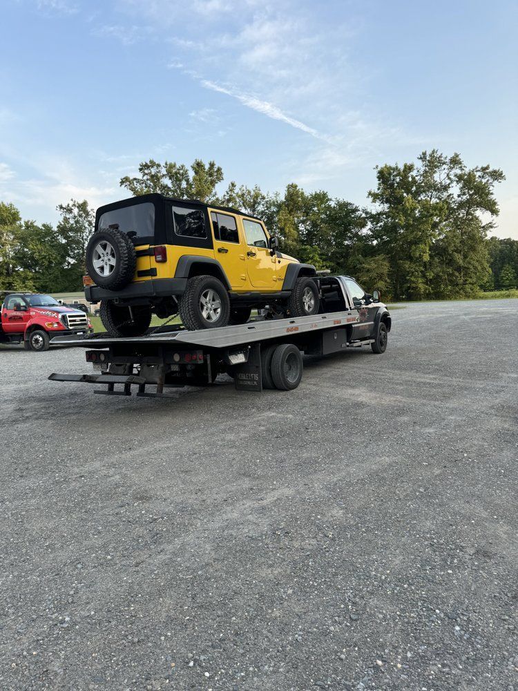 Yellow Jeep on a flatbed tow truck in a gravel lot, with trees and a red car in the background