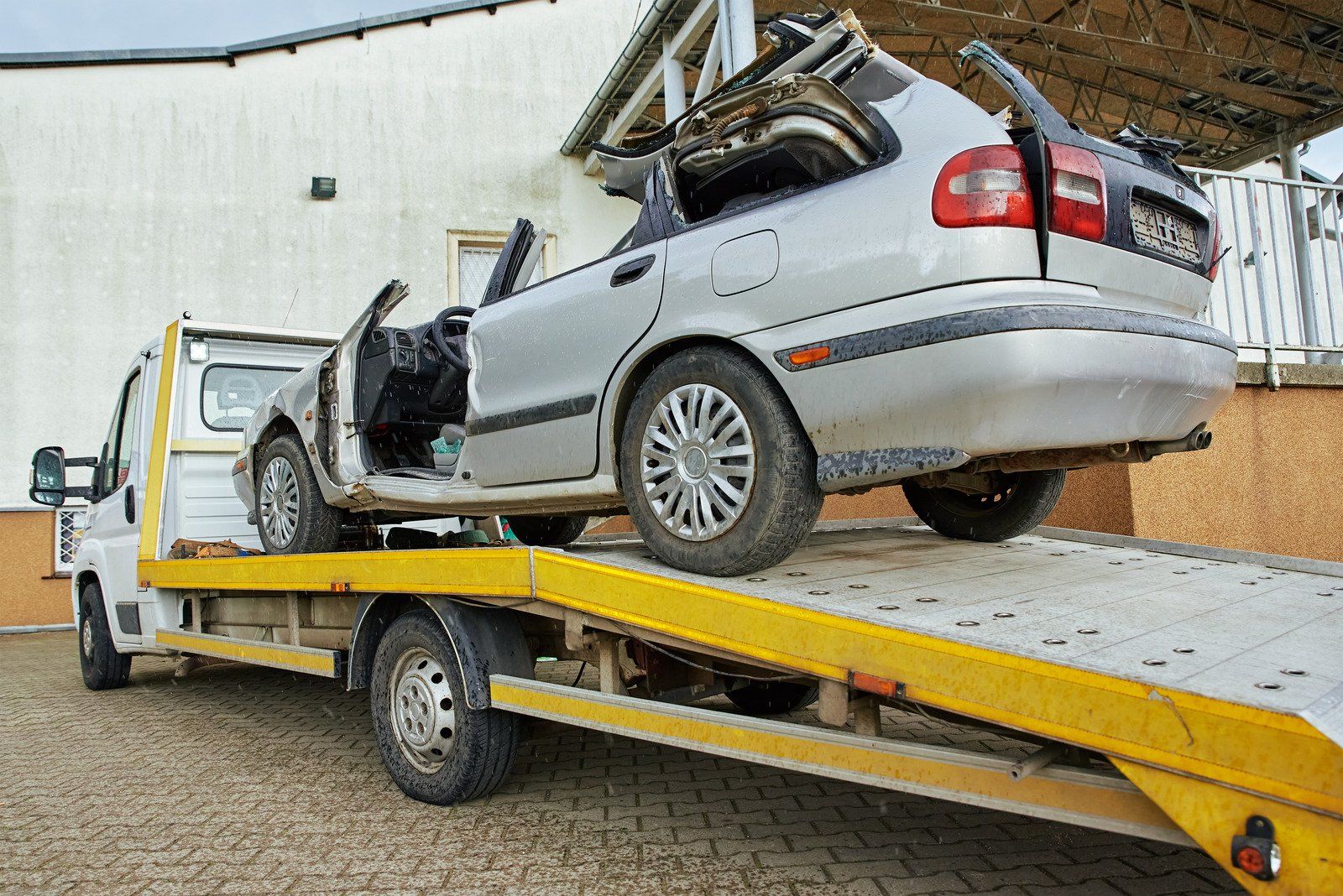 A severely damaged silver car on a flatbed tow truck. The car has missing doors and a collapsed roof.