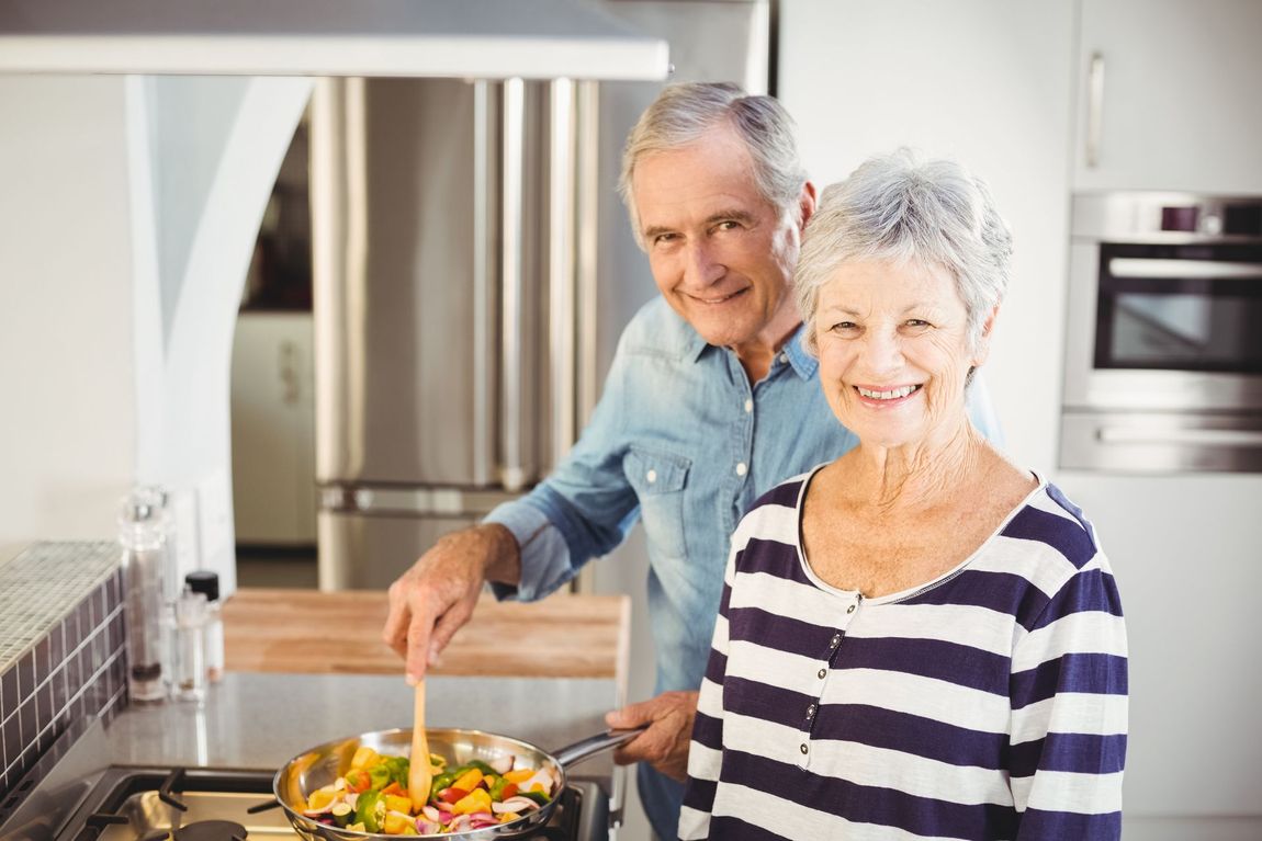 Two people cooking together in a bright kitchen, smiling beside a pan of vegetables.