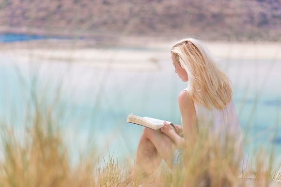 Person sitting by a turquoise lake, reading a book among tall grasses