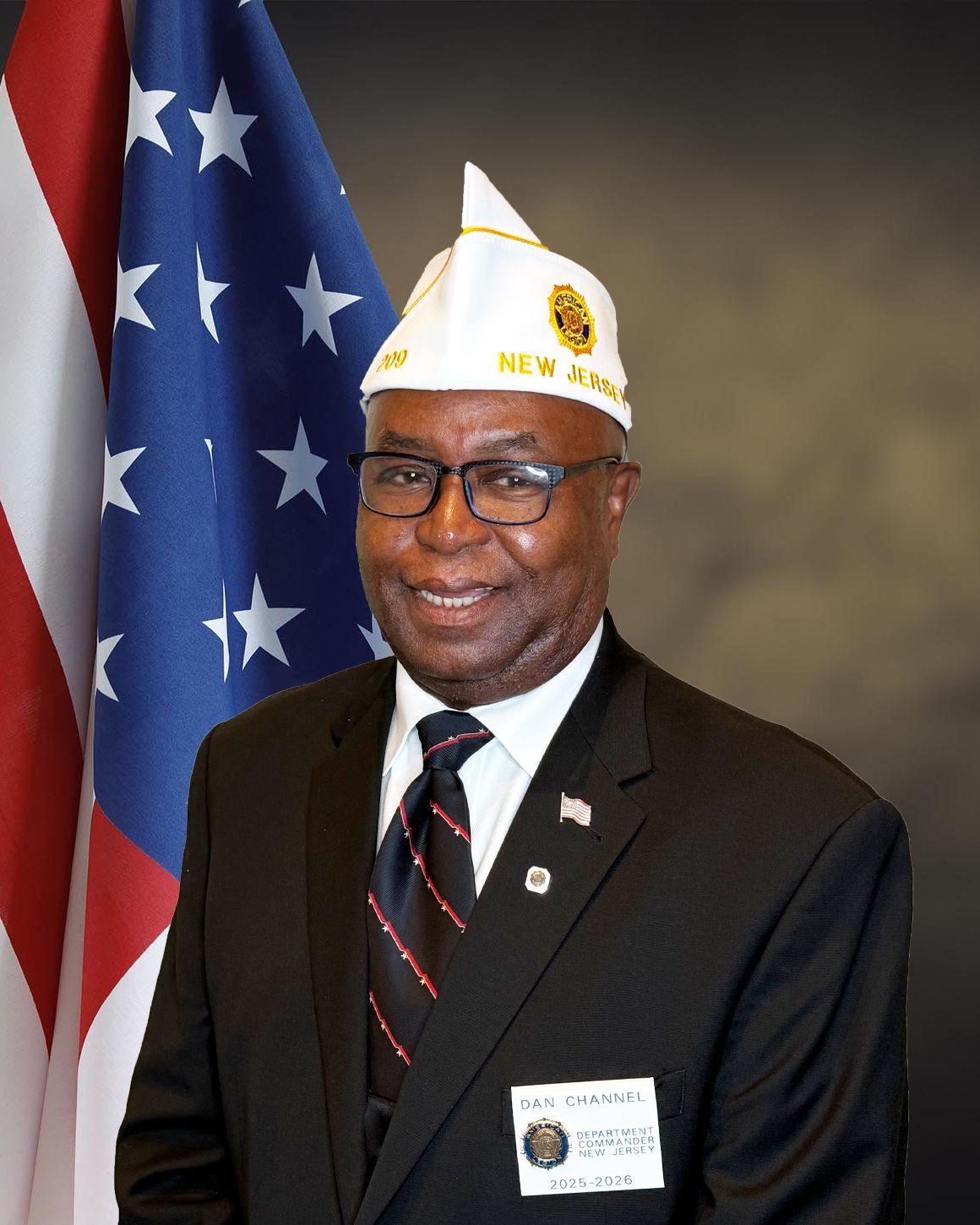 Man in suit and veteran's cap stands before US flag.
