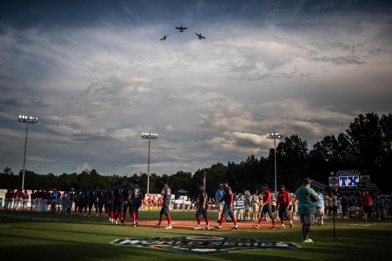Athletes on a baseball field look up as aircraft fly overhead during an outdoor event. Cloudy sky.