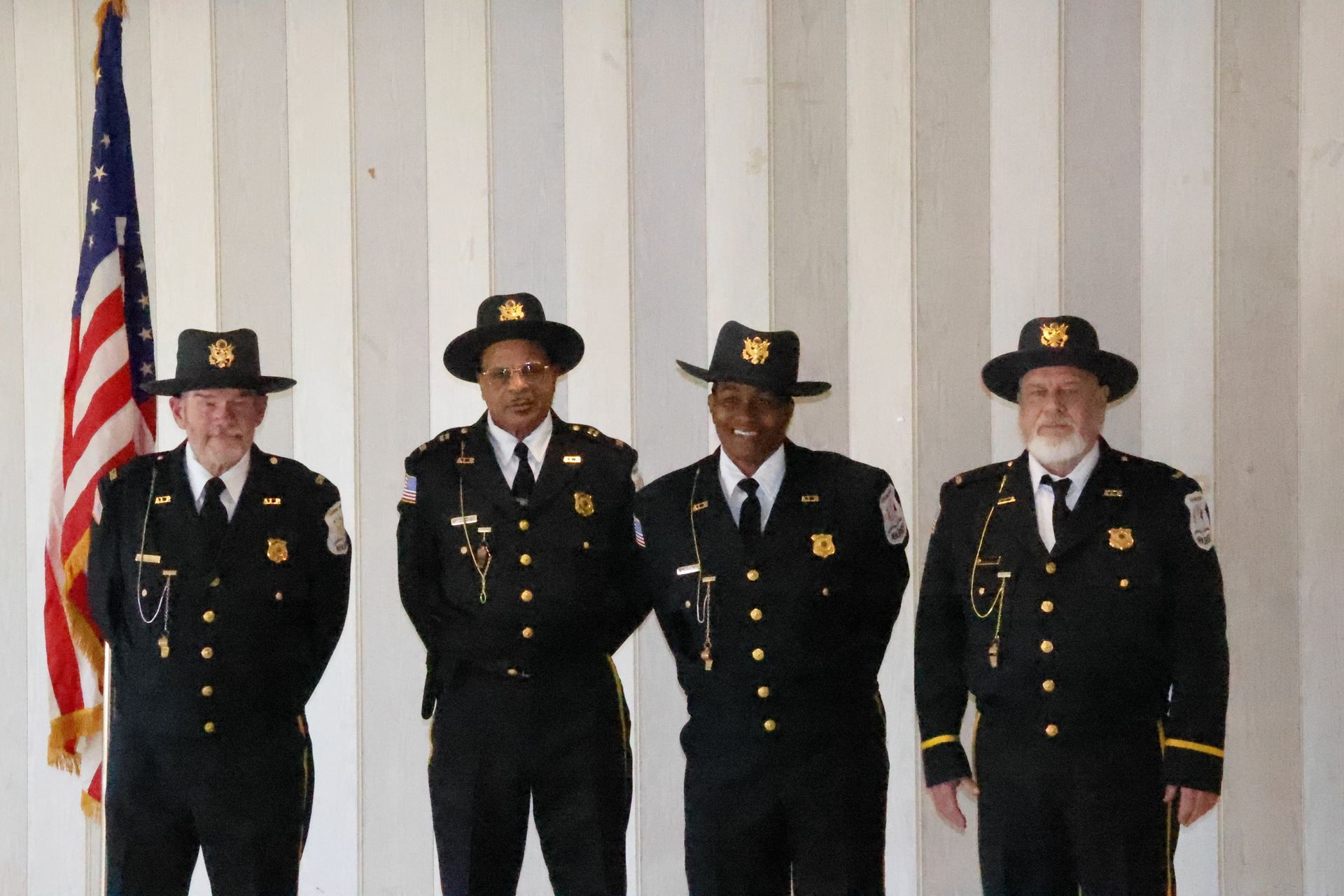 Four uniformed officers stand in front of a flag. They wear hats and dark suits, facing the camera.