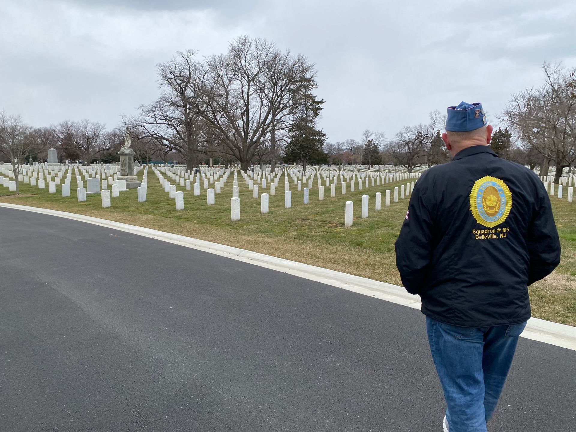 Man in blue hat and jacket walks past rows of white headstones in a cemetery on an overcast day.