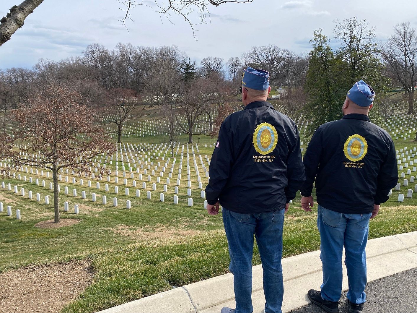 Two men in blue caps and jackets stand before rows of white headstones in a cemetery.